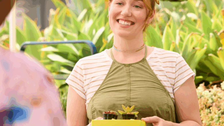 Eine lächelnde Person hält eine gelbe Schachtel mit zwei kleinen Sukkulenten in den Händen. Im Hintergrund sind weitere Pflanzen zu sehen.